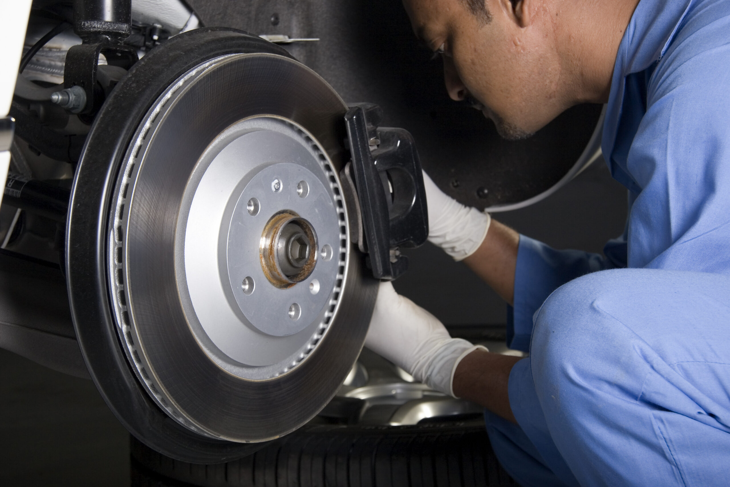 Mechanic in blue coveralls and white gloves working on a car brake system, focusing on the brake rotor and caliper.
