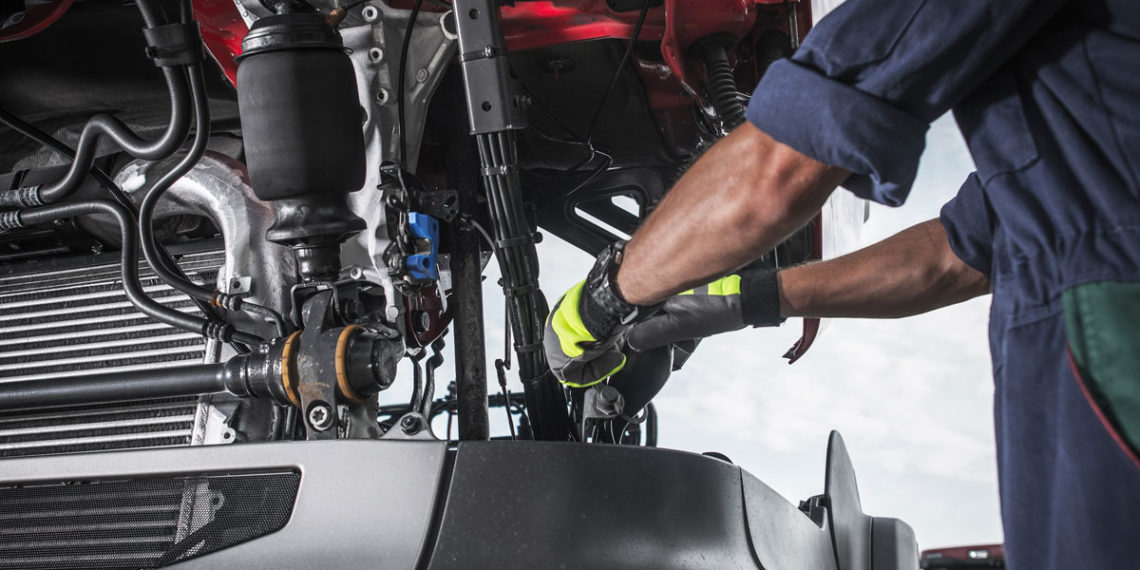 A mechanic wearing gloves repairs machinery under a vehicle hood.