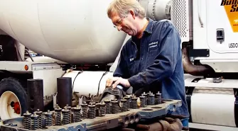 A mechanic works on an engine component beside a large vehicle, wearing coveralls and safety glasses, showcasing expert truck repair services.