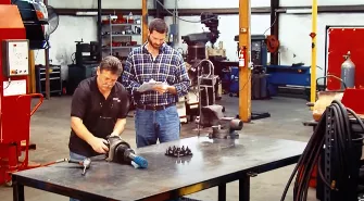 Two men working in a workshop: one with a tool in hand, and the other reading a document related to truck repair services. Industrial equipment surrounds them, emphasizing their expertise in commercial vehicle maintenance.